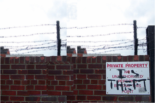 brick wall with barbed wire on top and a sign that has been grafittied to read Private Property is Theft