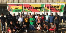 Group of people standing in front of a banner reading, "Free the land. Black August. Community Arts Festival"