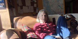 Heather Pritchard in a red sweater, seated on a brown couch with the afternoon sunlight streaming in.