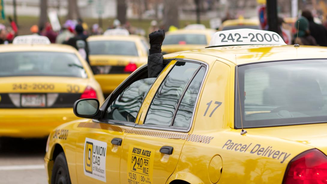 Line of Union Cabs at a protest. A driver is holding a raised fist out of their window.